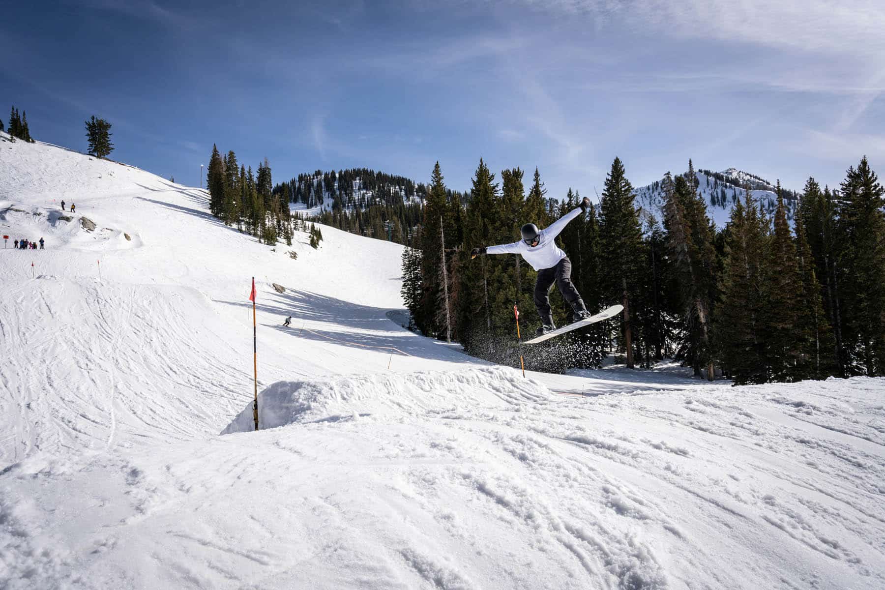 A snowboarder catches air in a daring mid-flight trick above a snowy ramp, with a backdrop of an audience and pine trees under the expansive blue sky, capturing the thrill and skill of snowboarding.