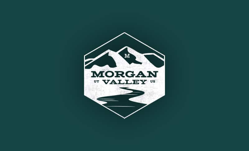 A hexagonal logo featuring a stylized depiction of snow-capped mountains and a flowing river, with ‘MORGAN VALLEY UT US’ prominently displayed, set against a dark green background with a rustic, distressed texture.